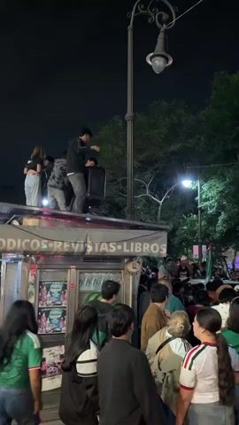 A nighttime crowd gathers around a newsstand, some people standing on top of the newsstand, holding a speaker. The crowd appears to be celebrating, with many wearing green shirts and holding Mexican flags. The newsstand has signs that read "PERIODICOS REVISTAS LIBROS" which translates to "NEWSPAPERS MAGAZINES BOOKS". The environment is outdoors with trees visible in the background and a streetlamp providing light.