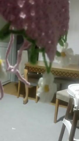 The video shows a close-up of a crocheted item, possibly a lampshade or a decorative hanging. The item is pink with a lacy pattern. Green crocheted vines with small white flowers hang from the bottom of the pink item. Some of the flowers have yellow centers. A pink crocheted strand is also visible hanging down. In the blurred background, a wooden dining table and chairs are visible, as is a portion of what appears to be a baby high chair. The lighting suggests the video was taken indoors.