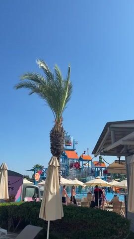 A water park scene unfolds under a clear, bright blue sky, suggesting a sunny day, likely midday. In the foreground, a large, beige beach umbrella casts a shadow, its fabric slightly wrinkled. To the left, the trunk and fronds of a tall palm tree dominate the frame, its leaves swaying gently. Behind the umbrella, a white food truck with pink accents and text that appears to read "L O S S" is partially visible.

The main focus of the background is a vibrant water park structure. It features blue and orange platforms, water slides, and a large, bucket-shaped water dispenser at the top, which is about to tip its contents. Several beach umbrellas are scattered around a shallow pool area where people are relaxing and children are playing in the water. A few individuals are visible enjoying the water park attractions. The overall atmosphere suggests a leisurely and fun day at a resort or aquatic facility. The location is identified as Trachoni, Cyprus.