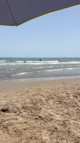 The video opens with a low-angle shot of a sandy beach, with the edge of a large umbrella visible in the upper left corner of the frame. The umbrella is dark gray with a yellow trim. The sand in the foreground is textured and appears dry. Beyond the sand, there is a calm ocean with gentle waves. Several people are visible in the water, some of whom appear to be swimming or wading. The sky is clear and blue. The camera remains stationary throughout the short clip.