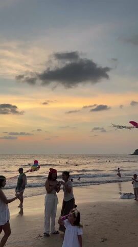 The video captures a serene beach scene at sunset. Several people are present, including adults and children. In the foreground, two women are standing close together, possibly looking at a phone or device. A child is blowing bubbles nearby. Towards the right, a man and a boy are standing on the sand, observing the scene. Other people are swimming or playing in the ocean. The sky is a blend of orange and grey, indicating the setting sun, and a few clouds are scattered across the horizon. Two kites are flying in the sky. The overall atmosphere is relaxed and peaceful, capturing a moment of leisure and family time at the beach.