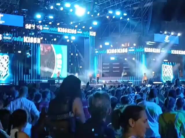 The video captures a lively outdoor event at night, likely a concert or festival, with a large crowd gathered. The stage is illuminated with bright blue lights and features large screens displaying graphics. In the foreground, the backs of many people are visible, some are holding phones up, presumably recording the event. In the midground, the crowd is dense and appears to be enjoying themselves. In the background, there are buildings with lit windows and signs, indicating an urban setting. Some people are standing on scaffolding near the stage, possibly part of the event crew. The overall atmosphere is energetic and festive.