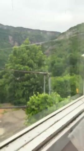 The video shows a view from a window overlooking a green, mountainous landscape. In the foreground, there are trees, some pipes, and a fence. The sky is overcast. The camera pans slightly to the right, revealing more of the mountains and vegetation. There are no people visible in the video. The scene appears to be rural or semi-rural.