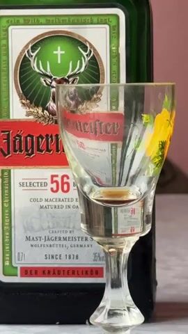 This is a close-up video of a bottle of Jägermeister liquor and a shot glass with a small amount of the liquor in it. The bottle is dark green with a label that features a deer head with antlers, a cross, and the text "Jägermeister" in red. The label also states "SELECTED 56 BOTANICALS," "COLD MACERATED ELIXIR," "MATURED IN OAK," "CRAFTED BY MAST-JÄGERMEISTER SE," "WOLFENBÜTTEL, GERMANY," "SINCE 1878," and "35% VOL." The shot glass is clear and has a small base, a stem, and a wide bowl. The glass has a yellow floral pattern on the side facing the camera. The background is blurred and appears to be a pinkish-brown surface. The video is very short, lasting only a few seconds, and shows no people or any interactions. The lighting is bright and highlights the label and the liquid.