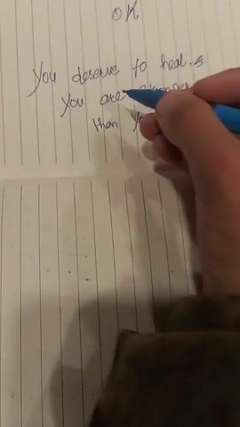 The video shows a close-up of a hand writing in a notebook with blue ink. The notebook has horizontal lines. The handwriting reads "You deserve to heal. You are stronger than you think". The word "OK" is written above this message, and part of another word is visible above "OK". The hand is holding a pen, and the sleeve of a dark, camouflage-patterned jacket is visible at the bottom of the frame.