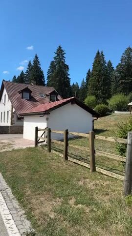 The video shows a side view of a white building with a red tile roof, set against a backdrop of green trees and a blue sky. The building appears to be a house or a small establishment, with several windows and dormers visible on the main structure. A smaller, white annex with a similar red roof is attached to the side of the main building. A wooden fence with wire mesh runs along the foreground, separating a grassy area from a paved path or driveway. The overall scene suggests a rural or semi-rural setting on a sunny day.