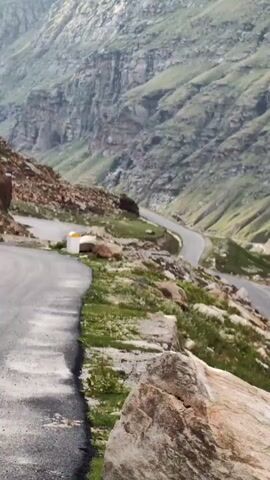 This video shows a winding road through a mountainous valley. The road is paved and appears to be in good condition. There are rocks and green vegetation on the sides of the road, and steep, layered cliffs rise up on either side of the valley. In the distance, there are fields and more mountains. The lighting suggests it is daytime.