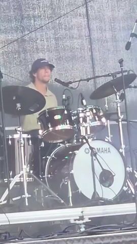 A man wearing a baseball cap and a short-sleeved shirt is playing a drum set on a stage. The drum set has a Yamaha bass drum. There are microphones set up around the drum set. The background appears to be a stage backdrop with some lighting.