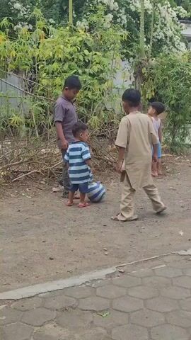 A group of young boys are playing soccer on a dirt path. The boy on the left is wearing a striped shirt and shorts, and is barefoot. He is facing the boy in the center, who is wearing a purple shirt and gray pants. The boy in the center is about to kick the ball. To the right, another boy in a tan outfit is watching them. In the background, there are trees, bushes, and a building. The ground is covered in dirt and some scattered debris. The sky is overcast.