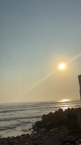 The video shows a beach scene with the sun shining brightly over the ocean. The sky is a light blue color.  The waves are gently rolling towards the shore, and there are rocks and debris along the shoreline. The sun's rays reflect off the water.  The scene appears peaceful and serene. There is a concrete post in the bottom left of the frame. There appears to be a structure on the right edge of the frame.