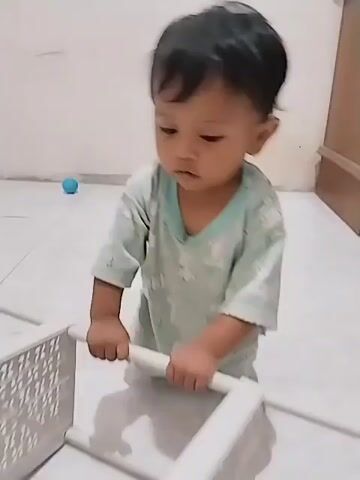 A young child, appearing to be around one to two years old, is kneeling on a tiled floor. The child is wearing a light green t-shirt with a faint pattern and light-colored pants. The child is holding onto a white, modular storage unit that appears to be made of plastic. The storage unit is composed of several cubby-like sections and horizontal bars. The child is focused on manipulating the storage unit. In the background, there is a wall and a corner of a room, with some objects visible, including what appears to be a child's tent with a cartoon character (Doraemon) on it, and a red object which may be a toy. The lighting suggests an indoor setting, possibly a home.