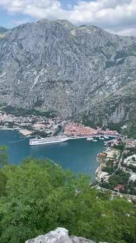 This is a vertical, medium shot video of the Bay of Kotor in Montenegro, taken from a high vantage point overlooking the water. In the foreground, lush green foliage partially obscures the view, with a gray, rocky surface visible at the very bottom. The main subject is the bay itself, a calm expanse of deep blue water.

Dominating the mid-ground is a large, white cruise ship docked near the shore. Beyond the ship, the town of Kotor unfolds, a picturesque settlement characterized by its terracotta-roofed buildings clustered tightly together. The town is nestled at the foot of a towering, rugged mountain range that fills the background. The mountains are imposing and stony, with patches of vegetation clinging to their slopes. The sky above is a mix of blue and white clouds, suggesting fair weather.

The activity taking place is static, depicting a scenic view of a coastal town and its bay, with a cruise ship present. The time of day appears to be daytime, likely late morning or early afternoon, given the bright sunlight and cloud cover. The overall mood is one of tranquility and natural beauty, with the grandeur of the mountains contrasting with the human settlement and the vastness of the sea. No visible text is present in the video.