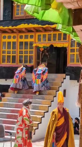 Two individuals in colorful robes with ornate headdresses are walking up a set of stairs towards a building with yellow-trimmed windows and green banners hanging overhead. In the foreground, two other individuals are visible. One is in a red and white robe with a dark head covering, and the other is in a yellow and maroon robe with a yellow hat. The stairs are painted with red and white stripes. The building appears to be made of dark stone.