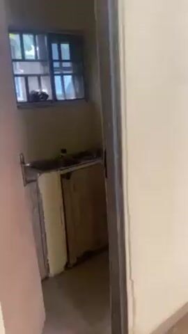 The video presents a small, simple kitchen area. A stainless steel sink is set into a yellowish countertop with a wooden cabinet below. A barred window is above the sink. The walls are painted a light yellow. The room is dimly lit. The video is taken from the doorway, looking in. There are some items on the countertop next to the sink. The floor appears to be tiled.