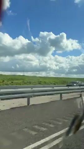 The video shows a view from inside a moving vehicle, likely a car or bus, traveling along a highway. The road is wet, possibly from recent rain. On the side of the road is a guardrail, and beyond that, a grassy field stretches towards the horizon. The sky is mostly blue with fluffy white clouds.  A truck passes on the highway in the opposite direction. The camera occasionally catches glimpses of the vehicle's interior or obstructions on the windshield, such as a partially visible red object.