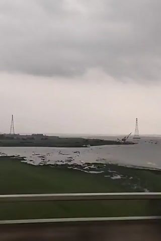 The camera moves along a bridge on a rainy day.  In the distance, two tall structures can be seen over a body of water.  Patches of green vegetation are visible on the left side of the screen.  The sky is overcast.  Raindrops streak the window from which the video is being recorded.