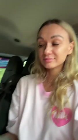 A blonde woman in a light pink shirt sits in the driver's seat of a vehicle and eats a single strand of pasta. She is holding the strand with her left hand and puts one end in her mouth, then pulls the rest of the strand into her mouth. Her shirt has a pink heart on it with "MILK" written across it. There is a tattoo on her left forearm. The vehicle's interior, including a headrest and air vent, is visible in the background. The woman looks directly at the camera as she eats.