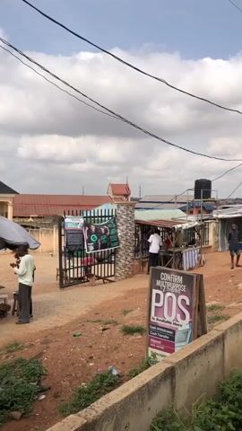 The video shows an outdoor scene in what appears to be a market or street corner in an African community. The ground is predominantly red dirt. There's a concrete drainage ditch in the foreground with some vegetation growing from it. Several small businesses or stalls are visible, constructed from corrugated metal and wood, with individuals standing and interacting around them. A group of people are gathered under a large, open umbrella. Several signs advertise local businesses, including a POS (Point of Sale) service for cash withdrawals and transfers, and a travel and freight company. Power lines crisscross overhead against a partly cloudy sky. Buildings with various roof types, including tiled and corrugated metal, are visible in the background. People are walking around and seem engaged in purchasing or selling goods.