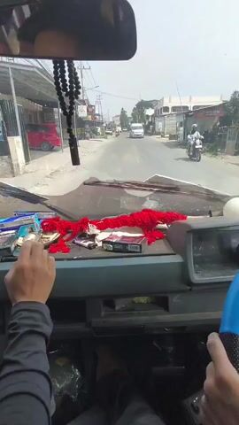The video shows the interior of a vehicle looking forward through the windshield. A person's hands are visible. The left hand, wearing a black sleeve, holds a cigarette and is in the process of smoking it, with the cigarette lit. The dashboard of the vehicle is visible, cluttered with various items including a pack of cigarettes, a red fluffy object, and a black beaded necklace hanging from the rearview mirror. Outside the vehicle, a street scene unfolds with buildings, other vehicles, and pedestrians. The road is paved, and the weather appears to be daytime and possibly sunny. The perspective suggests the video is recorded from the passenger seat or from the driver's seat if the driver is not actively driving at that moment.