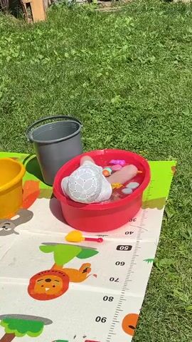 A young child, wearing a white sun hat with a gray pattern, is playing in a red basin filled with water. The child is mostly submerged in the water, with only their legs and feet visible above the surface. Various colorful toys, including what appear to be small plastic animals and blocks, are floating in the basin. A yellow toy shovel and a red toy rake are also present near the basin.

The basin is placed on a large, colorful mat with an animal theme, featuring a cartoon lion. The mat also has a ruler marking from 30 to 90 centimeters, suggesting it might be a height chart. To the left of the red basin is a gray bucket, and partially visible to the far left is a yellow bucket.

The scene is outdoors, with lush green grass covering the ground. In the background, there is a rustic wooden structure, possibly a shed or fence, and some fencing wires are visible. The lighting suggests it is a sunny day, likely midday given the strong shadows. The overall mood appears playful and relaxed, with the child enjoying their time in the water.

The visible text on the mat includes numbers indicating measurements: 30, 40, 50, 60, 70, 80, 90. There are also cartoon images of animals, including a lion. The location context is Азинский, Russia.