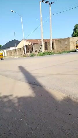Two yellow auto-rickshaws are driving on a dusty road. One auto-rickshaw is closer to the camera and is on the left side of the road. The other auto-rickshaw is further away and on the right side of the road. Both auto-rickshaws have passengers in them. There are buildings and power lines in the background. The sky is clear and blue.