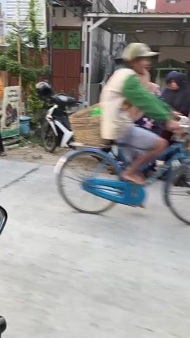 A woman in a pink hijab and a pink outfit rides a motorcycle loaded with baskets and bags through a street lined with buildings and trees. The woman is seen from behind. Two women in hijabs, one in blue and one in white, stand on the side of the street talking to a woman in a yellow shirt and blue pants. There are also motorcycles parked on the side of the street. The scene appears to be a residential or commercial area in Southeast Asia.
