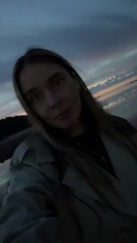 A young woman is standing outdoors in front of a body of water. The sky above is dark with streaks of orange and purple, suggesting it is dusk or dawn. The water reflects the sky and appears still. The woman is wearing a light-colored jacket over a dark top. Her hair is long and brown. She is looking at the camera and smiling slightly. The camera is held at a low angle, as if recording a selfie. The overall scene is peaceful and natural.