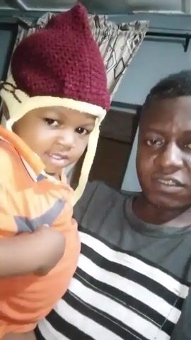 A man wearing a striped shirt is holding a small child. The child wears an orange shirt and a maroon and white knit hat. The child is looking at the camera and smiling slightly. The man holding the child is also looking at the camera, but appears more serious. The background shows a curtain with a geometric pattern.