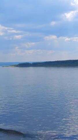 The video shows a wide, calm body of water under a partly cloudy blue sky. The water is a deep blue, reflecting the sky. On the horizon, there is a long, tree-covered shoreline. The camera appears to be moving slowly through the water, as evidenced by the slight blurring of the foreground on the left side of the frame. There are no people or specific objects visible in the scene. The environment is natural and serene.