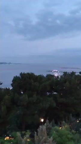 The video shows a panoramic view of a city skyline at dusk. The sky is cloudy and a deep blue, suggesting the transition from day to night. In the foreground, lush green trees and bushes obscure some of the view, with a lamppost visible among them. Beyond the trees, a body of water, likely a bay or sea, stretches out. On the water, there are several boats and structures.

In the distance, the city lights begin to twinkle as darkness falls. Prominent buildings, including a modern skyscraper with a curved design and what appears to be a stadium or large venue, are visible. The overall impression is of a bustling urban landscape by the water, seen from an elevated vantage point as evening descends. There are no people or overt actions depicted in the video, only a static landscape with changing light.