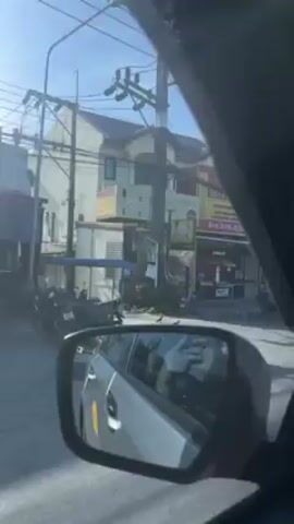 The video shows a street scene from the perspective of a car passenger. The car is moving, and the view is through the side window and side mirror. Several buildings with signage in Thai and English can be seen, including what appears to be a gold shop, a restaurant with the words "Pizza Pasta & More," and another restaurant. Cars, motorcycles, and utility poles are also visible. The sky is clear and blue.