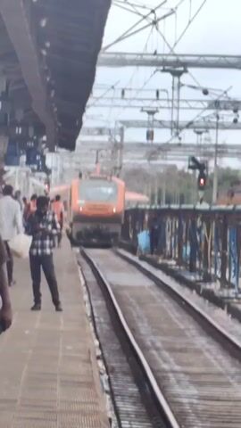 A train is approaching a station platform. A man is standing on the platform, looking at his phone. There are other people on the platform, some walking and some standing. The train is orange and white. There are overhead power lines and a signal light. The weather appears to be cloudy.
