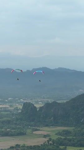 The video shows three paragliders with colorful wings flying against a hazy sky over a mountainous landscape. The paragliders are positioned at different distances and heights, with the closest one on the left. Below them, a valley is visible with green hills, rice paddies, and scattered trees. In the distance, several layers of mountains recede into the haze. The scene appears to be a scenic view from a high vantage point, likely captured during daylight hours with overcast skies.