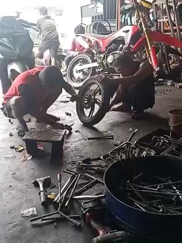 The video shows two men working on a motorcycle in a garage. One man, wearing a red shirt, is squatting and appears to be using a tool to work on the front wheel. The other man, wearing a black shirt, is also squatting and is holding the wheel of a red dirt bike. The garage is cluttered with tools, motorcycle parts, and other equipment. There are several other motorcycles in the background, including a white scooter. The floor is concrete and appears to be dusty. The lighting is dim, suggesting it might be indoors with artificial light.