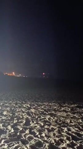 The video shows a nighttime beach scene with sand in the foreground and a dark sky. In the distance, there is a faint line of lights, possibly from a town or buildings. In the center of the frame, there are several bright pink flashes of light that appear to be fireworks. The flashes are intermittent and seem to be exploding in the sky above the distant lights. There are no people or distinct objects visible in the foreground or mid-ground. The overall impression is of a festive or celebratory event taking place on or near the coast.
