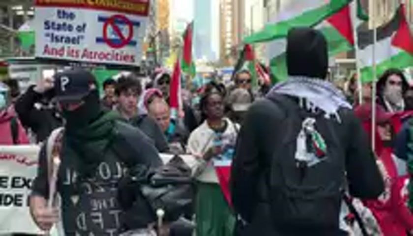 A daytime protest is underway on a city street, likely in New York, United States, with tall urban buildings and a clear blue sky forming the backdrop. A large crowd of people, including men and women, are actively participating in the demonstration.

Many individuals are waving numerous Palestinian flags, recognizable by their distinctive red, black, white, and green colors. Some protesters wear face coverings, such as black balaclavas or keffiyeh scarves. The crowd is energetically chanting slogans; "From the river to the sea, Palestine will be free!" and "Say it loud and say it clear!" are clearly audible.

Other visible objects include a person playing a drum with drumsticks. A large white protest banner with red lettering is partially visible, displaying the text "N THE STATE OF 'ISRAEL' AND ITS ALLIES." The atmosphere appears passionate and spirited, indicative of a strong collective sentiment.
