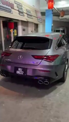 The video shows a dark grey Mercedes-Benz CLA 45 S Shooting Brake parked in what appears to be a garage or indoor parking area. The car is facing away from the camera, and the camera slowly moves from the rear of the car towards the driver's side. The car has tinted windows, and its taillights are illuminated. The license plate reads "RU 8822". In the background, there are signs for "Styling Garage," and other automotive-related services. There is also a shiny Airstream trailer in the background.  No people are visible in the shot.