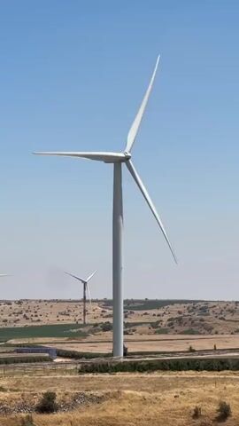 The video shows a wind turbine against a clear blue sky. The turbine has three white blades and a tall gray tower. In the background, another wind turbine with red and white markings is visible. The landscape surrounding the turbines appears dry and arid, with scattered vegetation and a dirt road or path in the foreground. The overall scene suggests a wind farm located in a rural or open area.
