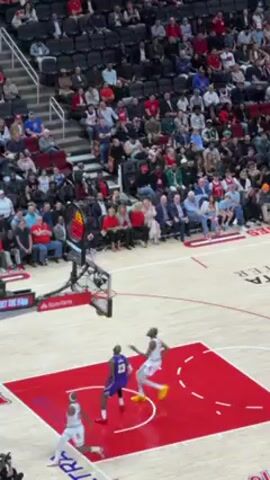 The video shows a basketball game in progress. The camera is positioned from an elevated angle, capturing a wide view of the court and a portion of the audience seated in the stands. LeBron James, wearing a purple Los Angeles Lakers jersey with the number 23, is the central figure. After a play near the basket, James turns and runs up the court, pointing his right index finger towards the ceiling. The court has a red and white design with the words "TOYOTA CENTER" and "HOMETOWN HEROES" visible. Other players in opposing white and red jerseys are visible, as are the crowd members who are mostly wearing casual clothing.