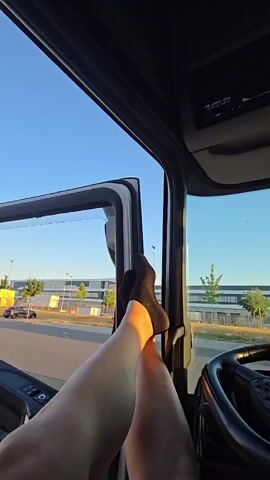 A first-person perspective video shows a person's legs and feet propped up in the open doorway of a truck cab. The person is wearing black socks. Outside the truck, the sky is clear and blue, and the sun is casting a warm glow. The background shows a paved area with some buildings and trees. A car is visible in the distance on the left. The steering wheel of the truck is visible on the right side of the frame. The video pans slightly upwards, revealing a portion of the truck's interior ceiling.