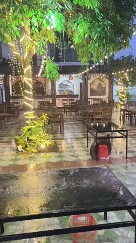 The video shows an outdoor seating area of a restaurant or cafe during a rain shower. The space is decorated with strings of yellow lights draped from trees and overhead. Several tables and chairs are visible, some of which are wet from the rain. In the foreground, a black table is covered in water droplets. A red plastic stool is placed next to a black metal table in the middle ground. The floor appears to be tiled and also wet. Trees with green foliage frame the scene. In the background, there is a building with decorative arches and what appears to be a bar or counter area. The overall atmosphere is calm and somewhat deserted due to the weather.