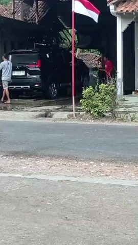 A black SUV is parked under a covered structure. A person wearing a red shirt is spraying the side of the SUV with a hose. Another person wearing a light-colored shirt is standing near the back of the SUV. A red and white flag is flying on a pole in front of the SUV. The ground is paved in the foreground and appears to be dirt and gravel in the background.