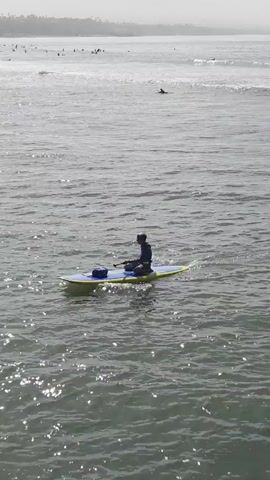 A person is paddleboarding on the ocean. The person is seated on the paddleboard and is using a paddle to move through the water. There are other people paddleboarding and surfing in the background. The water is choppy and there are waves. The sky is overcast.