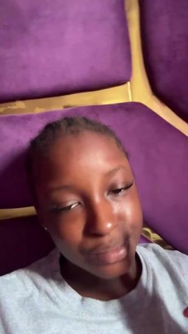 A young woman with brown skin, wearing long eyelashes, is positioned against a purple tufted armchair. She’s wearing a gray t-shirt and has her hair pulled back. She gives a thumbs up, and then holds one finger up. She appears to be speaking.