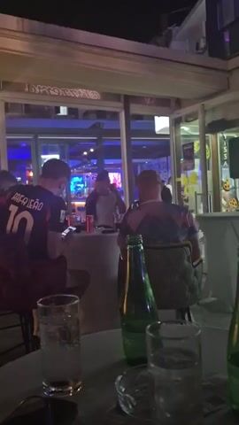 The video shows a dimly lit indoor setting, possibly a bar or restaurant, at night. In the foreground, two glasses filled with clear liquid and ice, along with two green glass bottles, are placed on a table. Further back, several people are visible. To the left, a person wearing a dark shirt with "LEAO 10" on the back is seated at a bar. In the middle, another person wearing a colorful, patterned shirt is seated. Towards the right and in the background, a bar area is visible with shelves of bottles. A large screen displays a sports game with green fields. Several high-top tables with white cloths are set up in the seating area. The atmosphere appears casual and social.