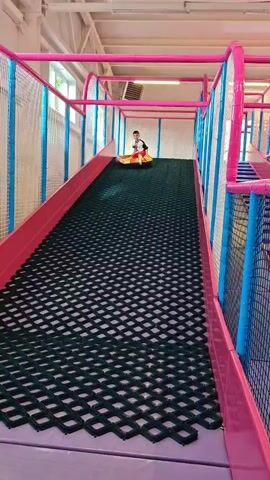 A young boy is sitting on an inflatable tube at the bottom of a slide in an indoor playground. He is wearing a white shirt with black sleeves, red shorts, and white socks. The slide has a pink border and a green textured surface. The playground appears to be brightly lit with natural light coming from windows in the background. The boy is smiling and looking at the camera. The inflatable tube has orange and yellow colors. There is another inflatable tube next to it. The scene conveys a sense of fun and play.