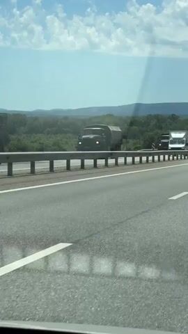 The video, filmed from a moving vehicle, captures a highway scene on a bright, sunny day. Initially, a dark green truck with a canvas-covered bed, possibly a military-style vehicle, is seen traveling in the opposite direction on the divided highway. A Russian license plate is visible on the front of this truck. Following it is a white truck, also moving away from the camera on the opposite side of the road, which bears some Russian text on its side, advertising a service with a phone number beginning with "+7 977 971 19 00".

As the video progresses, the camera shows the highway stretching forward, with several other cars and trucks visible in the distance in both directions. The road is asphalt with clear white lane markings and is divided by a metal guardrail. The background features green, tree-covered hills or mountains under a clear blue sky with scattered white clouds. A red and white striped road marker is briefly visible on the left side of the highway. The scene depicts a journey on a rural highway in what appears to be Saratovskaya, Russia.