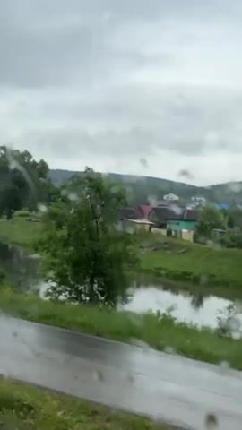 The video shows a view from a window with raindrops on it, looking out at a rural landscape. A river or stream runs through the scene, with lush green grass and trees lining its banks. In the distance, there are a few houses, suggesting a small village or settlement. The sky is overcast, indicating cloudy or rainy weather. The perspective suggests the video is being recorded from a moving vehicle, possibly a train or car, as the scene slowly pans to the right. There are no people or animals clearly visible in the frame. The overall mood is serene and a bit melancholic due to the weather conditions.
