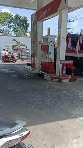 A video shows a gas station with a red and white pump labeled "1 BIO SOLAR". A blue truck with Indonesian flags is parked near the pump, laden with speakers and other items. In the foreground, a person is sitting on a motorcycle, facing away from the camera. Another person on a red scooter is visible in the background. The gas station has a covered canopy and appears to be in a rural or suburban setting with trees and buildings visible. The overall atmosphere seems to be bustling, possibly for an event or celebration.