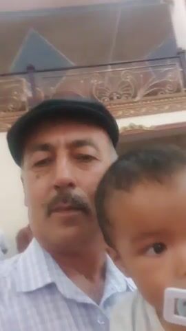 This media file captures a selfie of a man holding a young child. The man, wearing a dark cap and a light-colored striped shirt, has a mustache and appears to be in his 50s or 60s. He is looking directly at the camera with a neutral expression. The child, held in his arms, is a toddler with dark hair and is wearing a light-colored outfit and a pacifier. The child's gaze is directed slightly off-camera.

The background suggests an indoor event or gathering. In the upper portion of the frame, an ornate balcony with decorative metalwork is visible, hinting at a formal or traditional architectural style. Below this, strings of small, bright lights are strung across the space, creating a festive atmosphere. Other people are visible in the background, some blurred and indistinct, indicating a social occasion. The lighting appears to be artificial, suggesting it could be evening or an indoor venue. The overall mood conveyed is one of familial affection and a shared moment at an event.

There is no visible text in the media file. The location context of Rizamahalla, Uzbekistan, aligns with the visual cues of traditional architecture and the general appearance of the people.